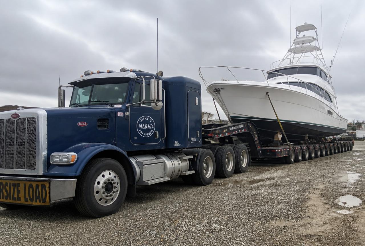 Navy heavy-haul truck with a multi-axle hydraulic trailer under a sportfishing yacht at a boatyard